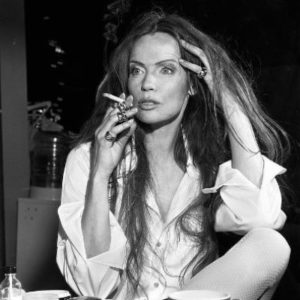 Veruschka I by Michel Comte, model sittin at a table in a white shirt, smoking