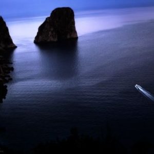 Capri Dreams by David Drebin, a boat on a quiet ocean in the dark next to rocky cliffs