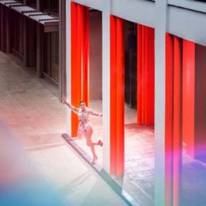 Fly with me by David Drebin, model in transparent dress leaning out of a building colored in purple and red light