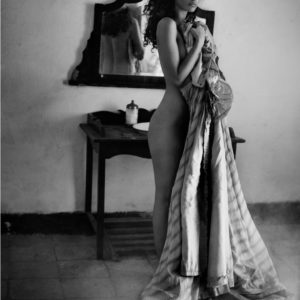 Sicily Palermo by Vincent Peters, a model with dark courls standing in front of a rustic vanity, nude, covering her body parsially with striped fabric